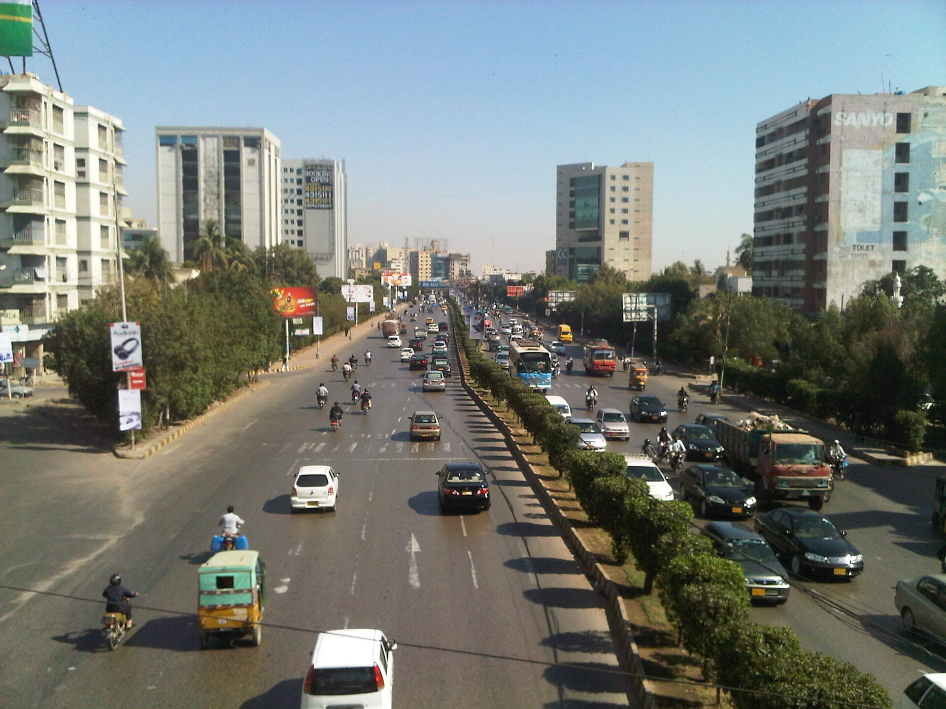 A wide, busy boulevard in Karachi lined with buildings and trees under a clear sky.