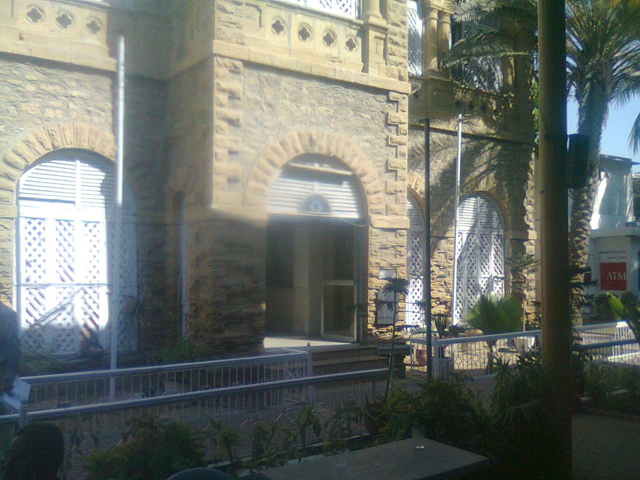 The stone facade and arched windows of the Karachi Press Club building, flanked by palm trees.