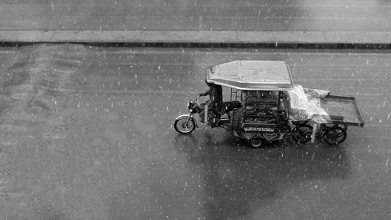 Black-and-white photograph of a Pakistani rickshaw on a wet road in heavy rain.