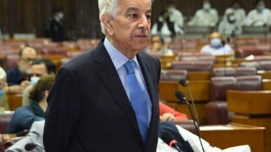 Defence Minister Khawaja Asif, wearing a dark suit and blue tie, speaks at a microphone in a legislative chamber with lawmakers seated behind him.