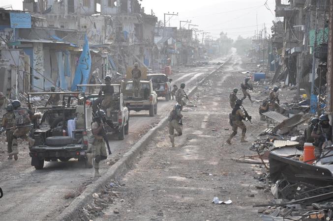 Pakistan Army personnel in combat gear moving along a rubble-strewn street lined with damaged buildings during a clearance operation.