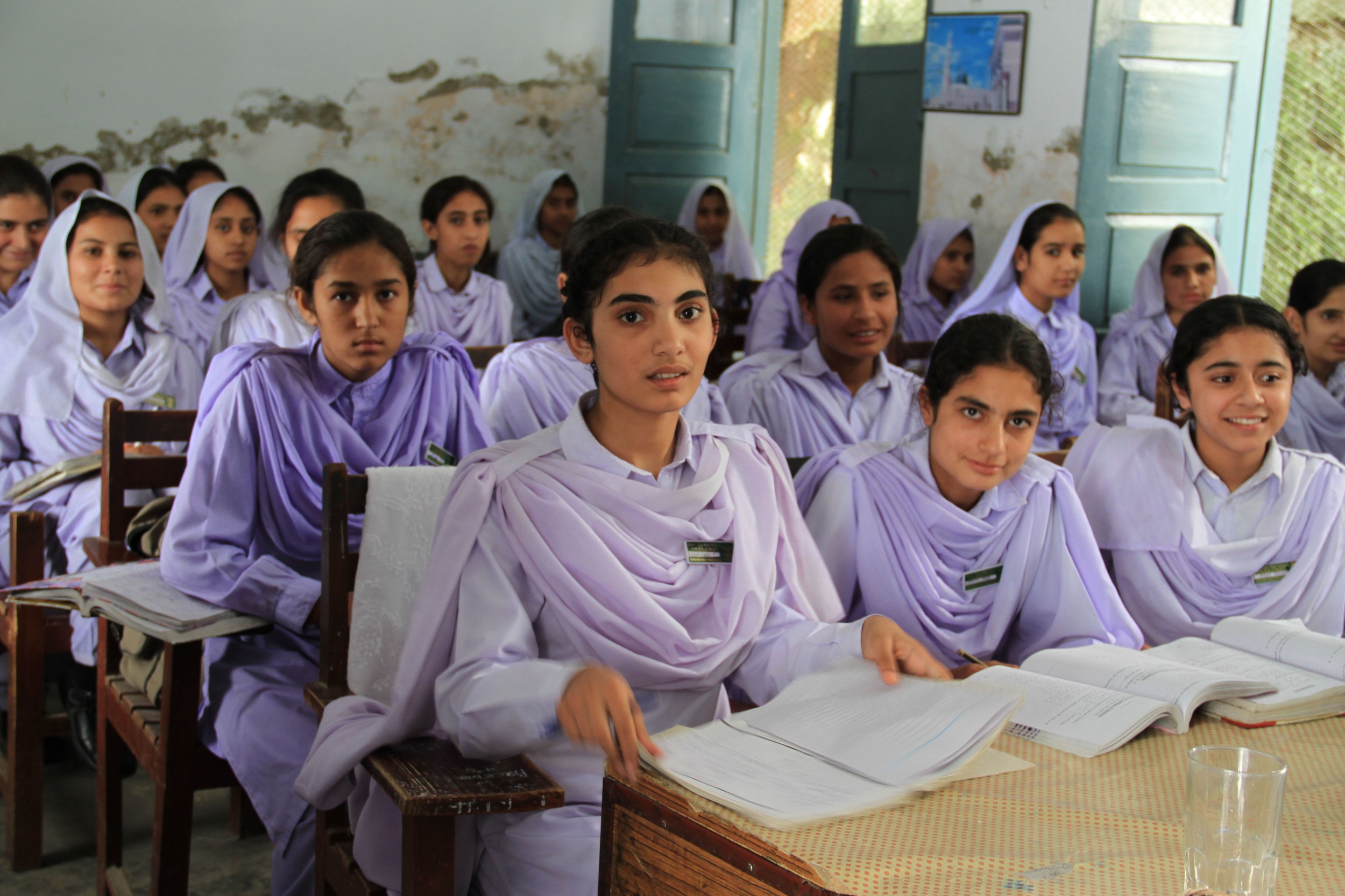 Schoolgirls in pink uniforms read textbooks at wooden desks inside a government primary-school classroom in Khyber Pakhtunkhwa, with light falling through the open doorway.