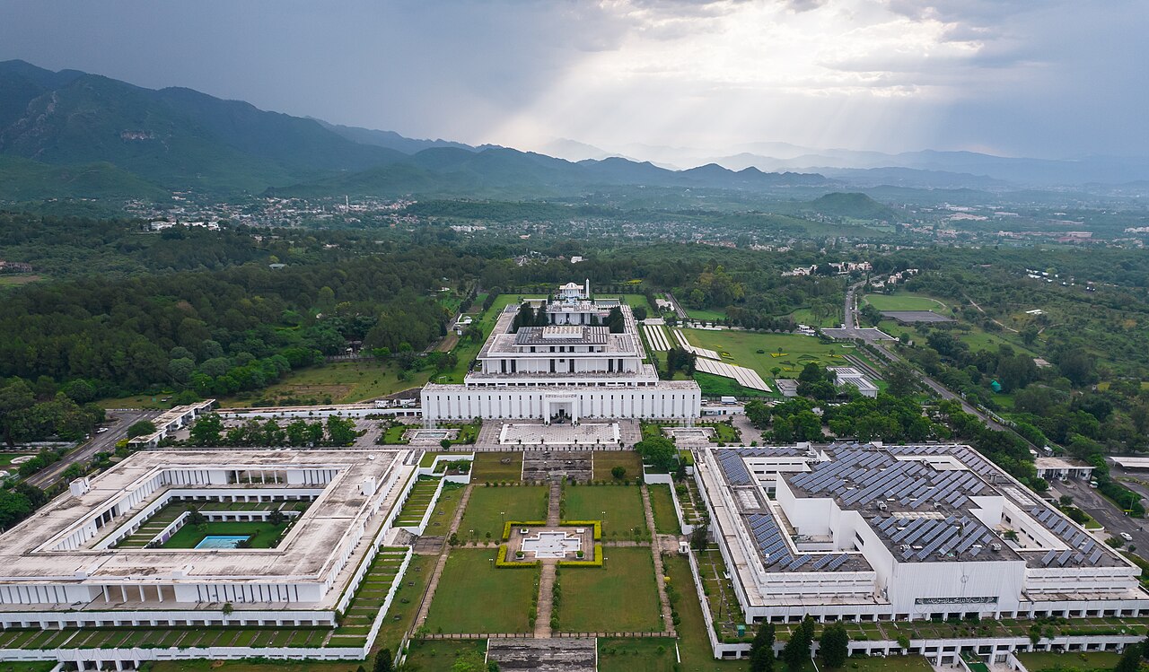 Aerial photograph of the white-domed Aiwan-e-Sadr presidential palace in Islamabad, with the surrounding gardens and Margalla Hills in the distance under a clear sky
