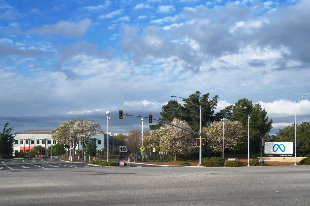 A white Meta sign with the company's blue infinity logo at the entrance to its Menlo Park headquarters, beside a tree-lined intersection