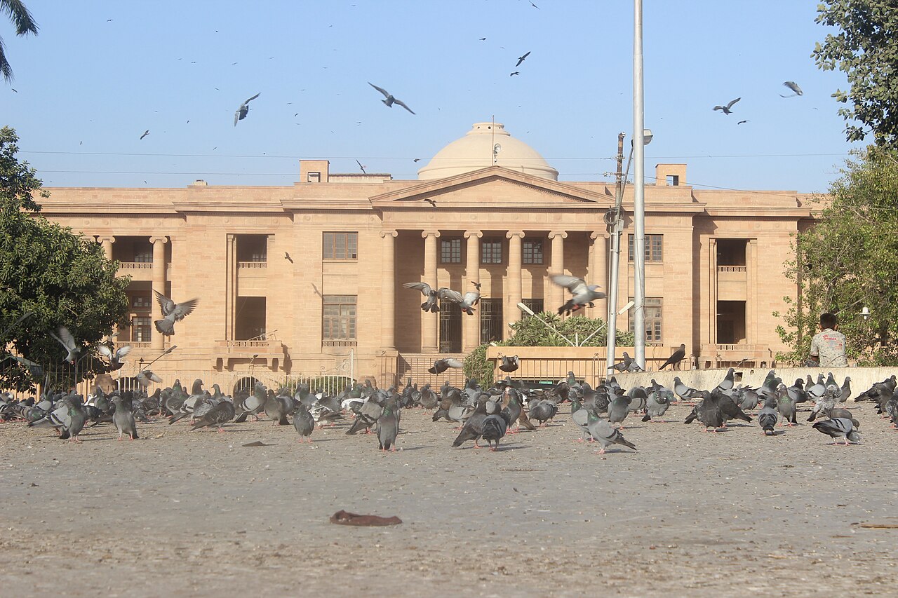 A sandstone court building with a central dome, columns and arched windows, seen across a paved courtyard with pigeons in the foreground.