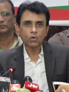 MQM-P chairman Khalid Maqbool Siddiqui speaking into a cluster of microphones at a media appearance, wearing a dark jacket over a light shirt.