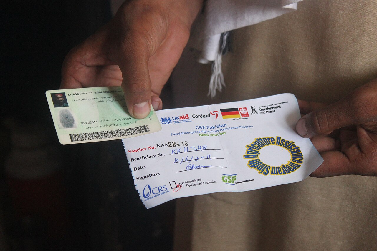 A pair of hands holds open a green Pakistani national identity card beside a printed seed voucher.