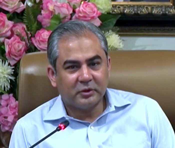 A man with short grey hair in a light blue button-down shirt speaks behind a black microphone, with pink and white floral arrangements behind him.