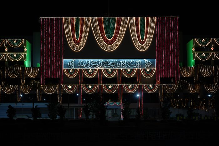 A large government building at night decorated with rows of small golden lights and red vertical light strands, with illuminated calligraphy on the central facade.
