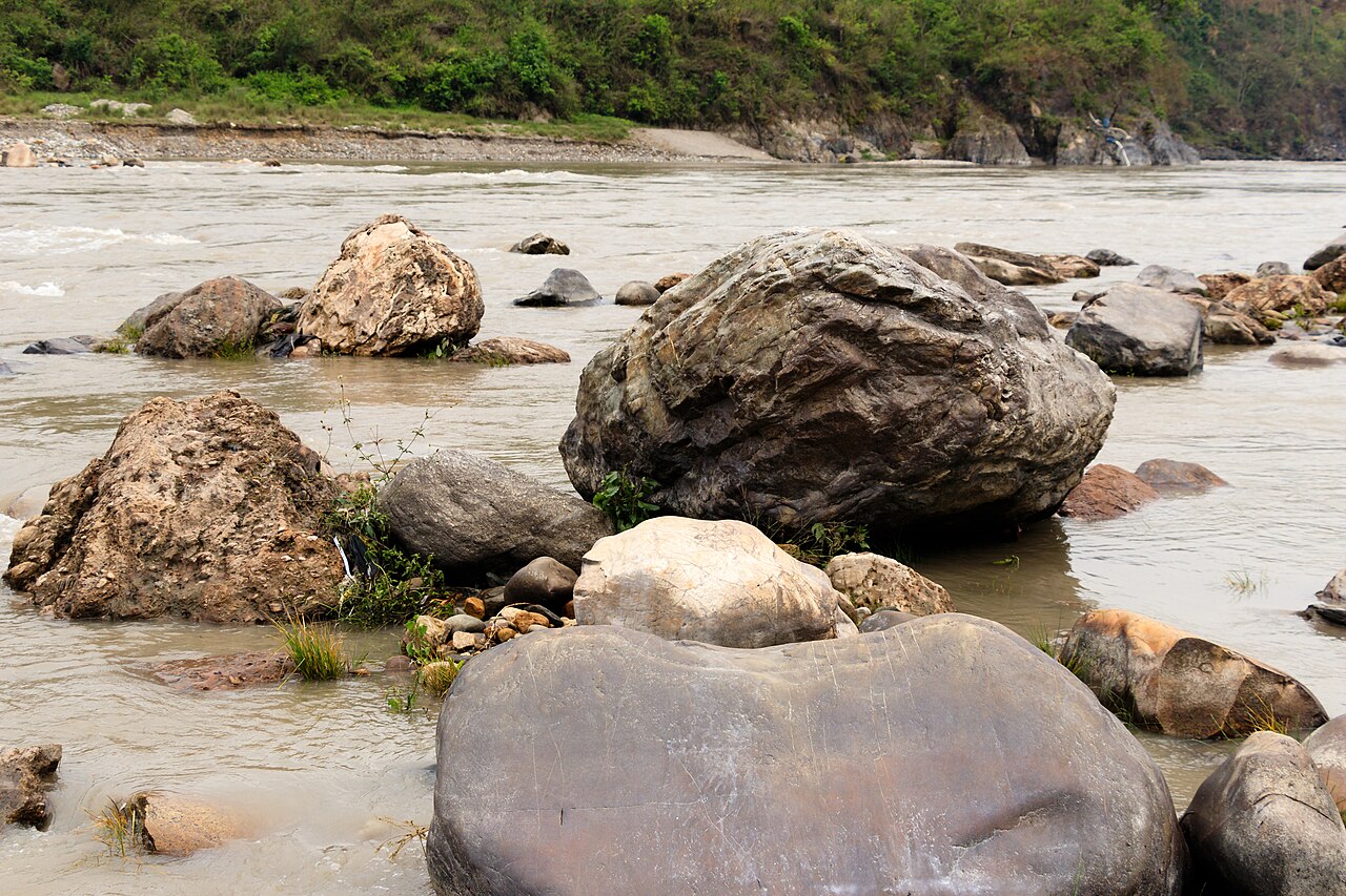 Large boulders in the Trishuli River with a wooded bank beyond, photographed in May 2019