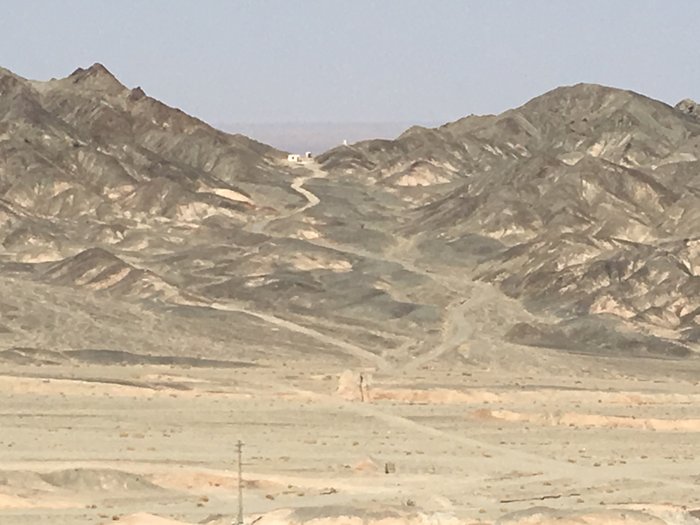 Arid, rocky mountains with a winding dirt track leading toward a small white building on a ridge, with a valley floor and transmission poles in the foreground near Saindak.