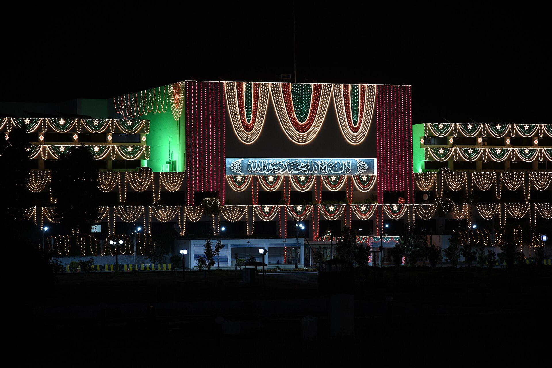 The Parliament House of Pakistan in Islamabad, a large modern building with a sloping roof, seen from an open paved area with flagpoles.