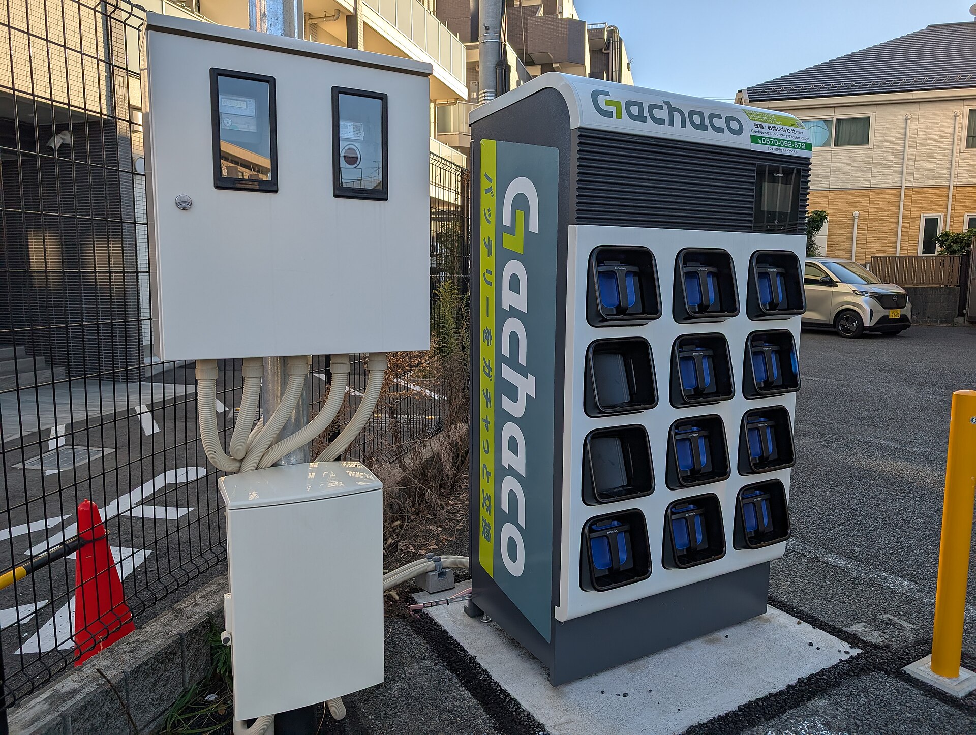 A row of battery-charging cabinets at a Japanese battery-swap station photographed at street level, with power units, cables and indicator lights visible behind glass panels.