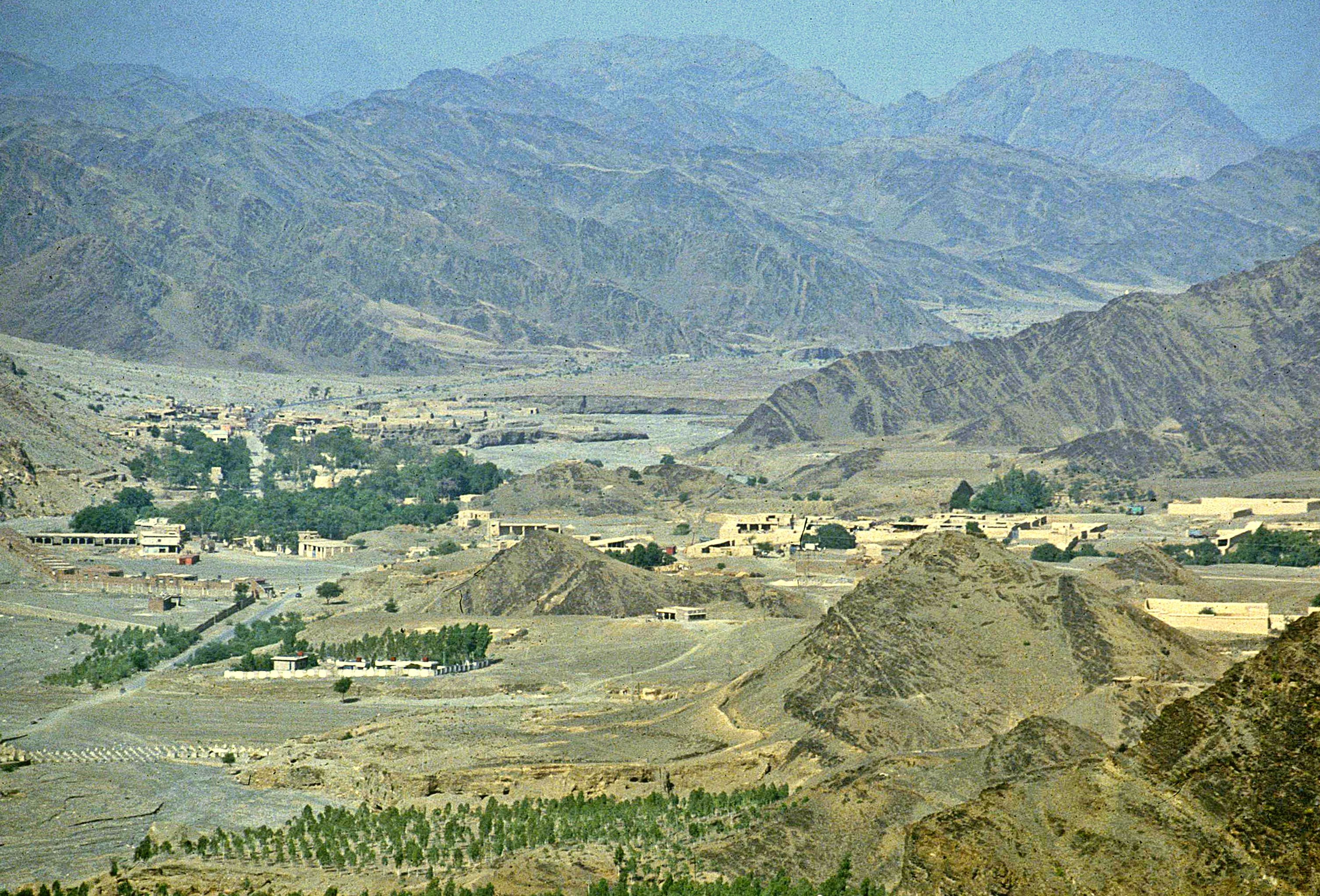 View from the Khyber Pass looking across the border toward Afghanistan, showing a settled valley of low buildings and cultivated terraces in arid, mountainous terrain.