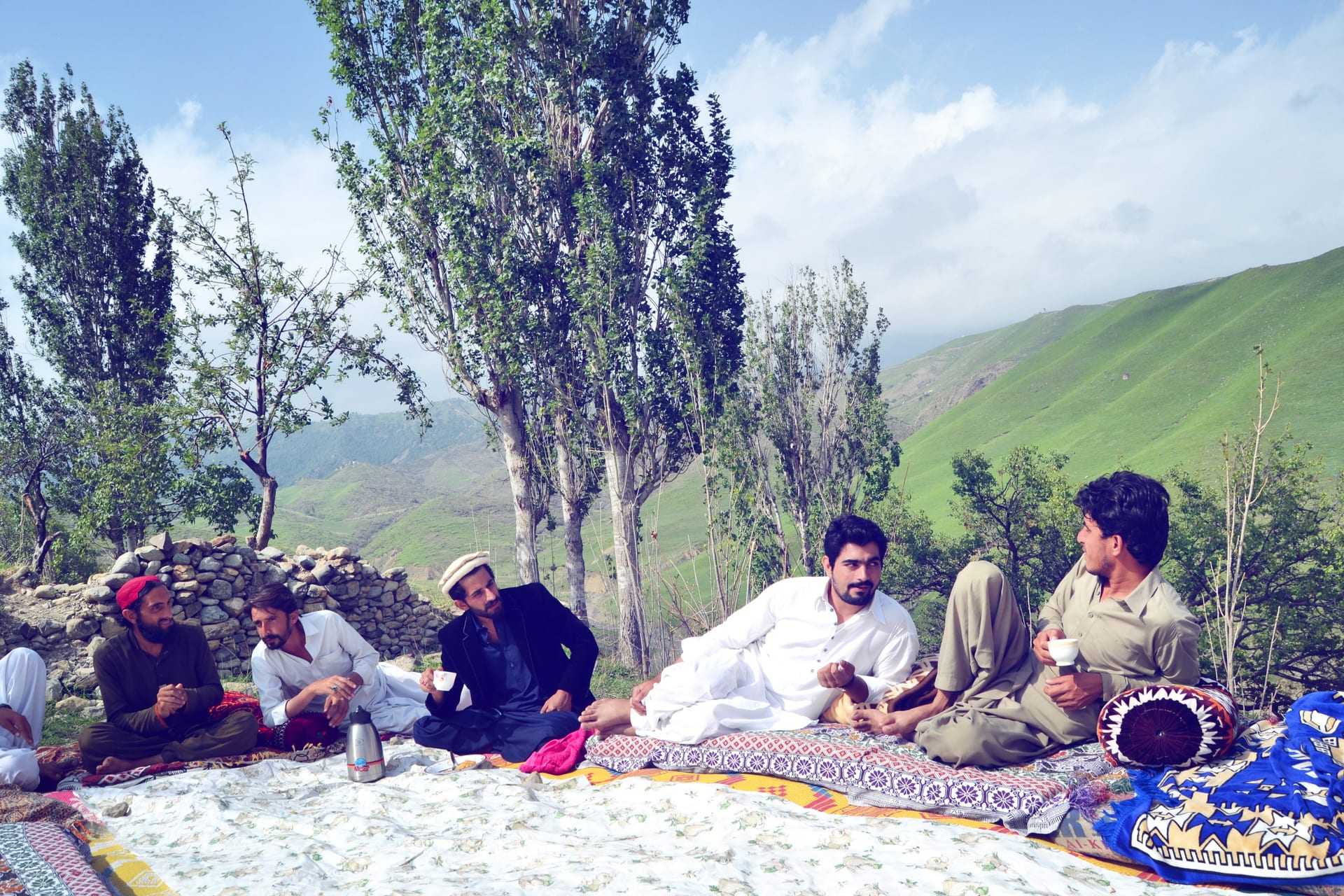 Five men in traditional dress sit on a charpai with tea cups and a metal kettle in front of a dry-stone wall, with terraced green hills and tall trees rising behind them in South Waziristan.