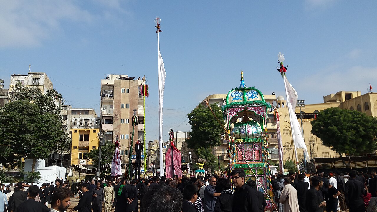 A Muharram mourning procession moves along a street in Karachi, with participants carrying banners and a decorated replica.