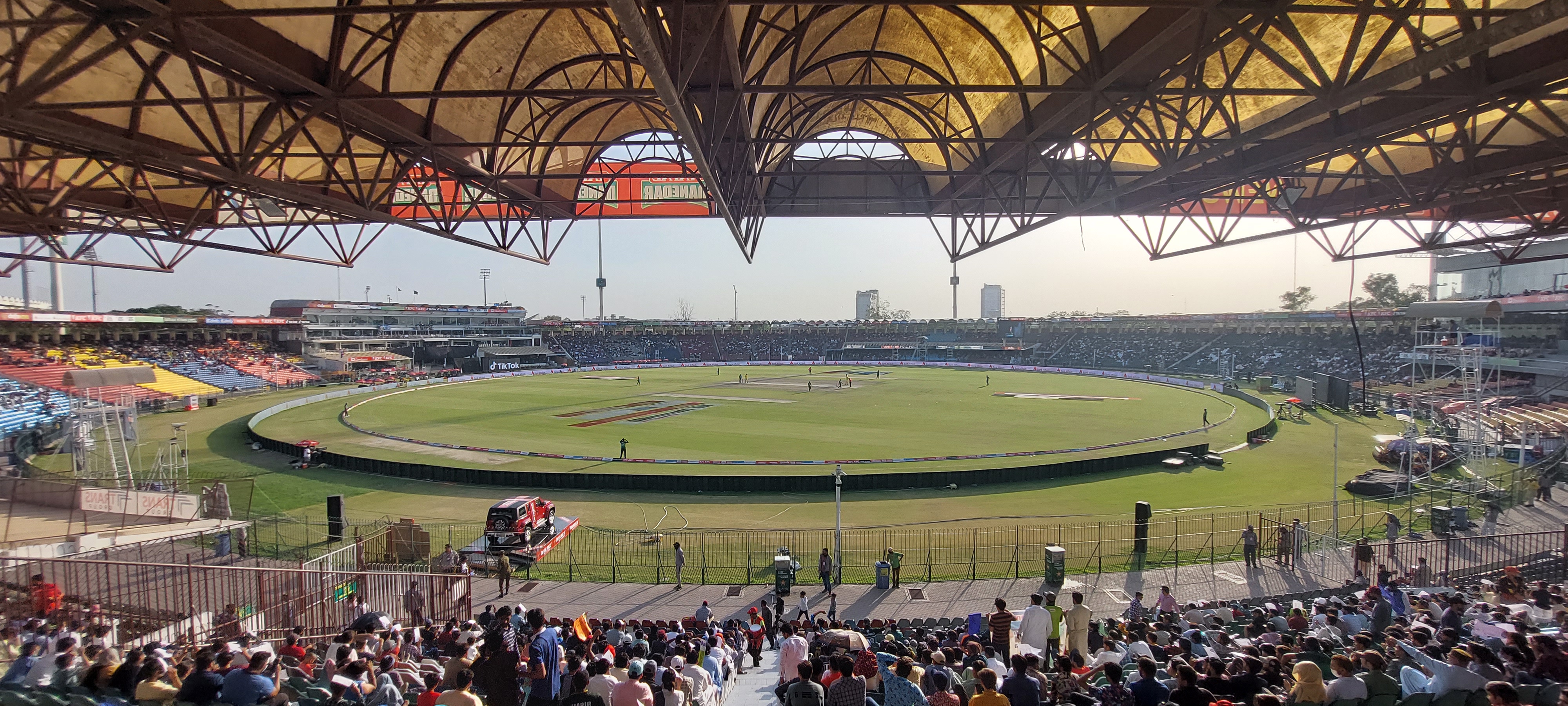 Floodlit cricket match in progress at Gaddafi Stadium, Lahore, with fielders spread across the ground and spectators in the stands
