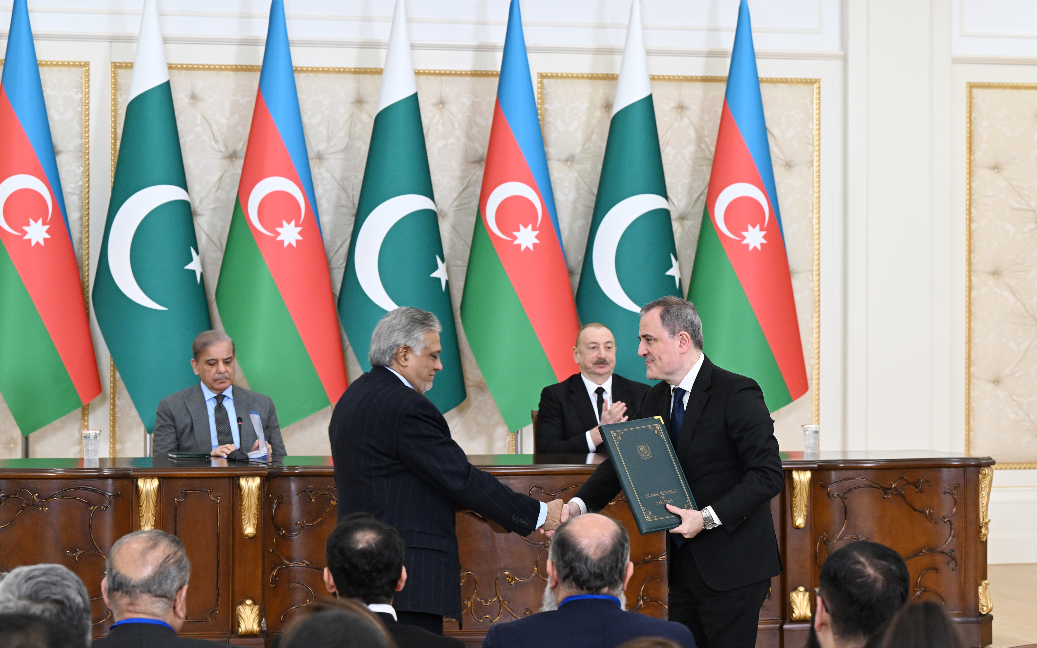 Two officials seated across a polished wooden table exchanging signed documents, with a man in a dark suit and a woman in a light blue jacket on one side and officials in dark suits and a woman in a dark patterned headscarf on the other, with small national flags in the background.