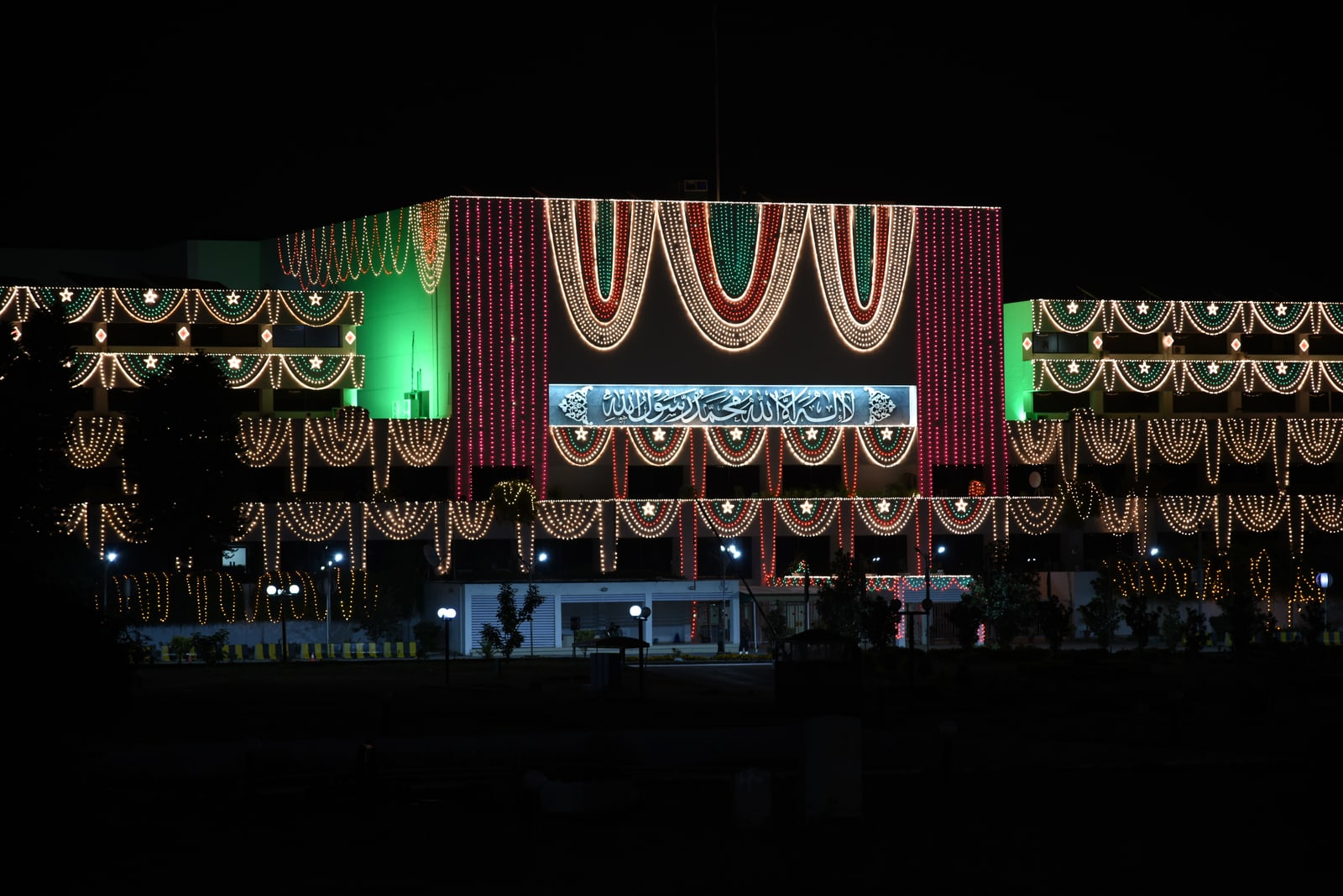Pakistan's Parliament House in Islamabad at night, its facade covered in strings of red, green and white lights around a central illuminated Urdu sign.