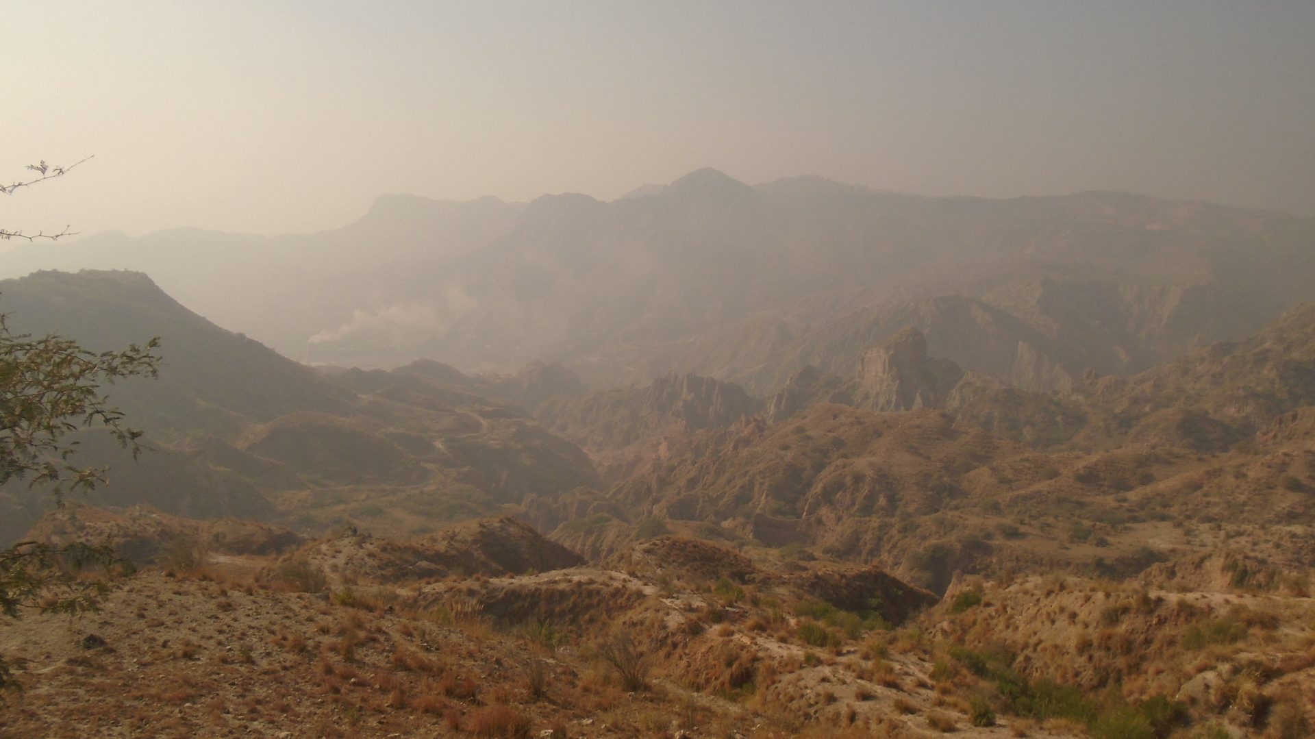 A wide, hazy view over the dry brown ridges of the Salt Range near Dandot in Punjab, with a cement plant and its chimneys faintly visible in the valley below and a thick dust haze hanging over the hills.