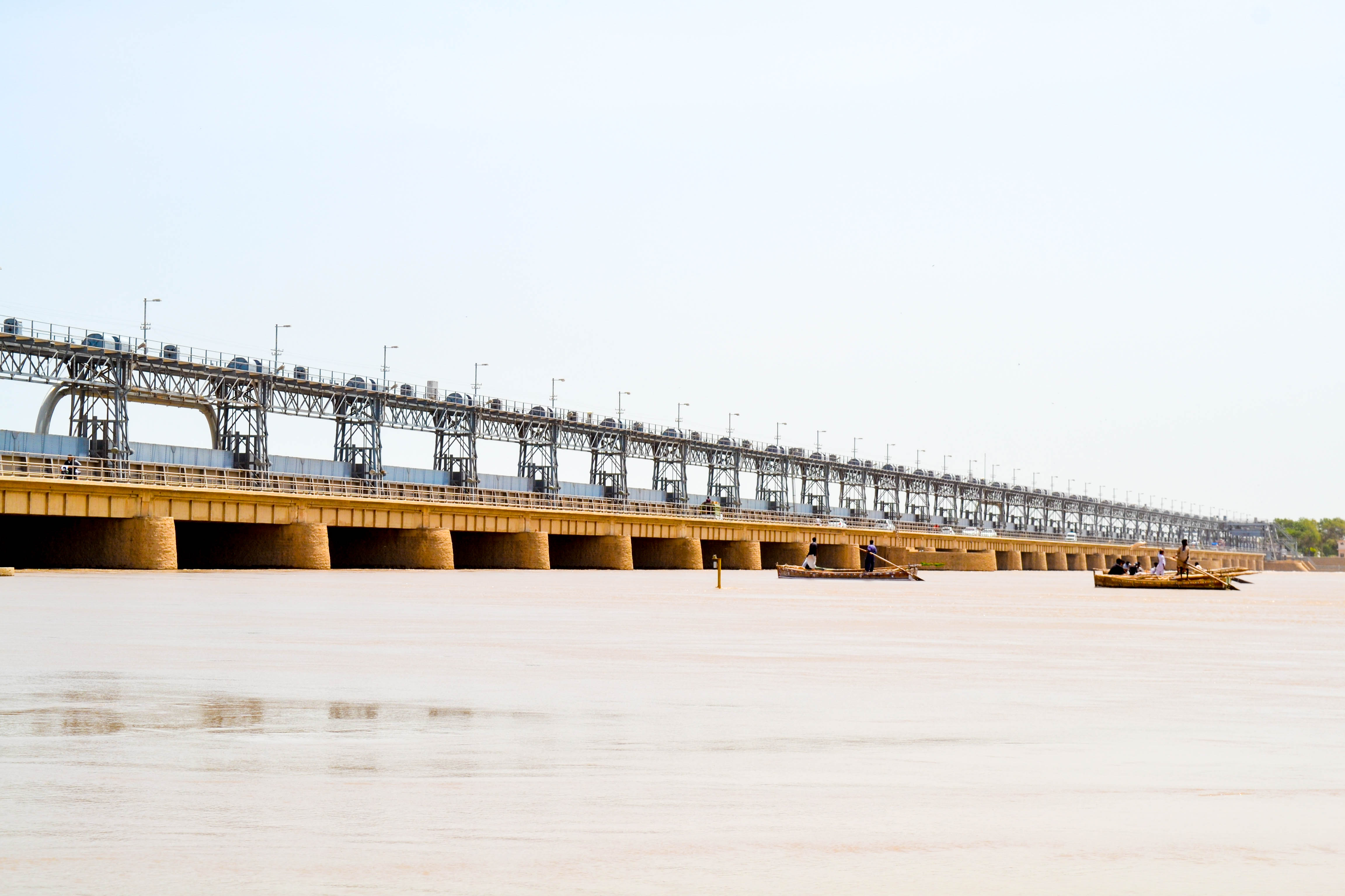 A broad river flows past a barrage structure, with small wooden fishing boats moored near the bank in the foreground.