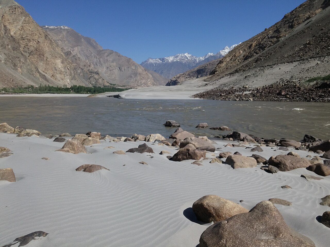 The Indus River flowing through a rocky valley near Skardu under a bright sky, with shallow water spreading over the riverbed in the foreground.