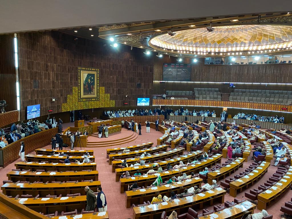 Wide view of the National Assembly chamber in Islamabad, with rows of wooden desks and maroon seats arranged in a horseshoe, a gilded domed ceiling and the electronic voting board above the members' gallery.