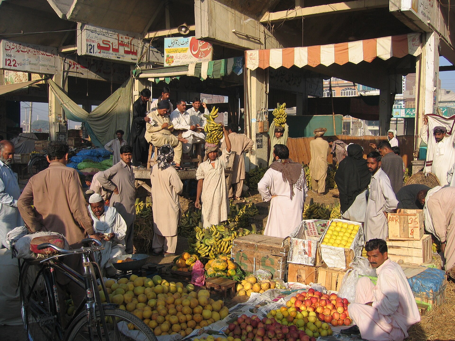 A crowded open-air fruit and vegetable market in Multan, with heaps of citrus and apples, hanging bunches of bananas, crates of green vegetables, vendors and shoppers, and Urdu signboards over the stalls.
