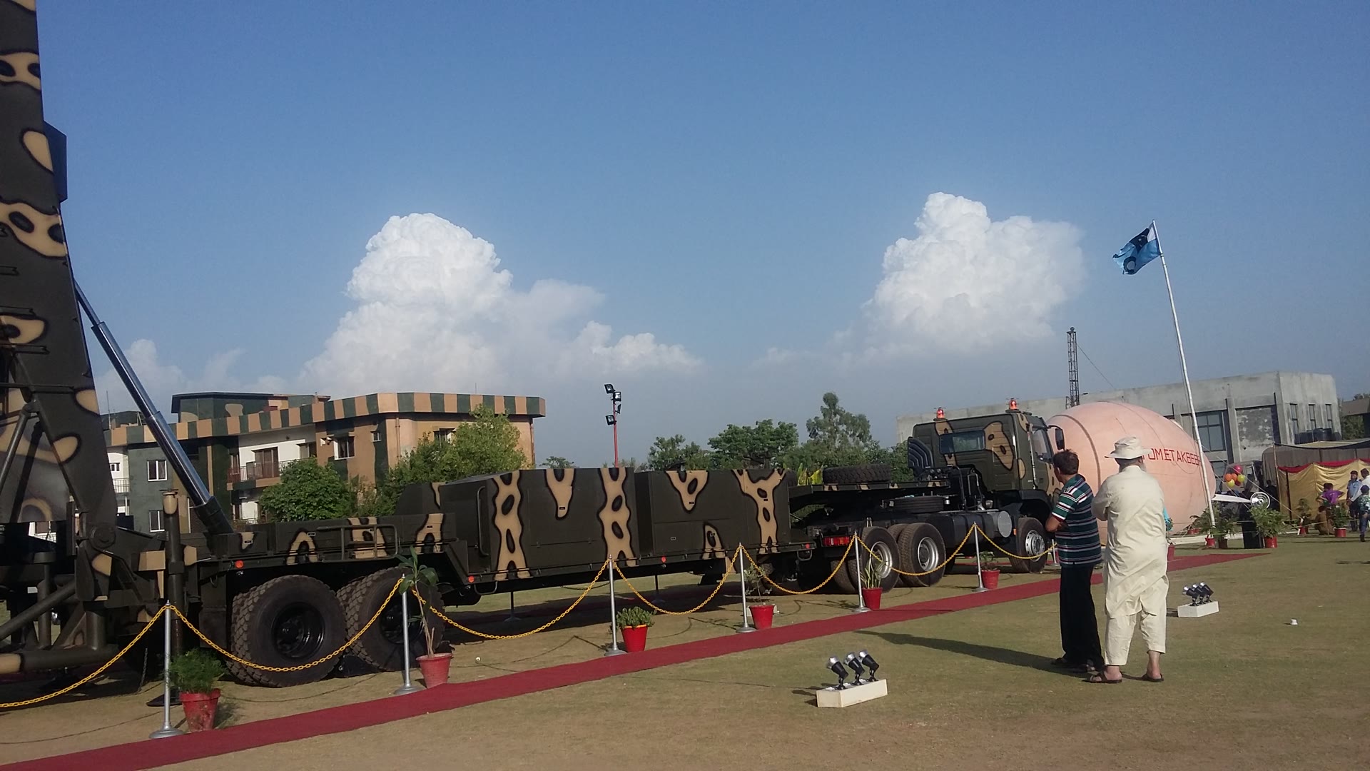 A camouflaged Pakistan Army transporter-erector-launcher and its flatbed trailer on display outdoors, with a Pakistani flag, spectators and buildings behind it.