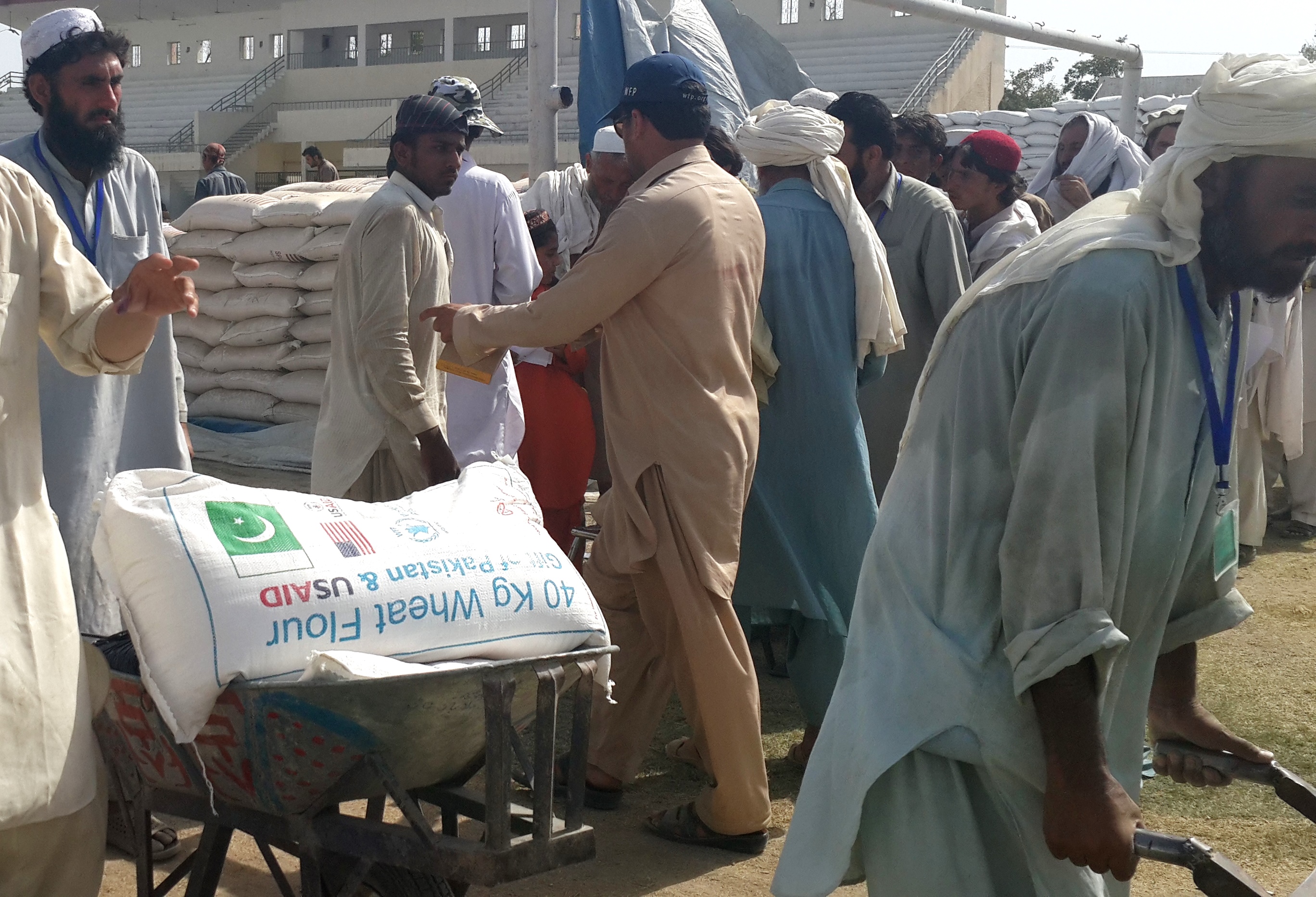 A crowd gathers at an outdoor food distribution point, with sacks of supplies and staff behind a counter.