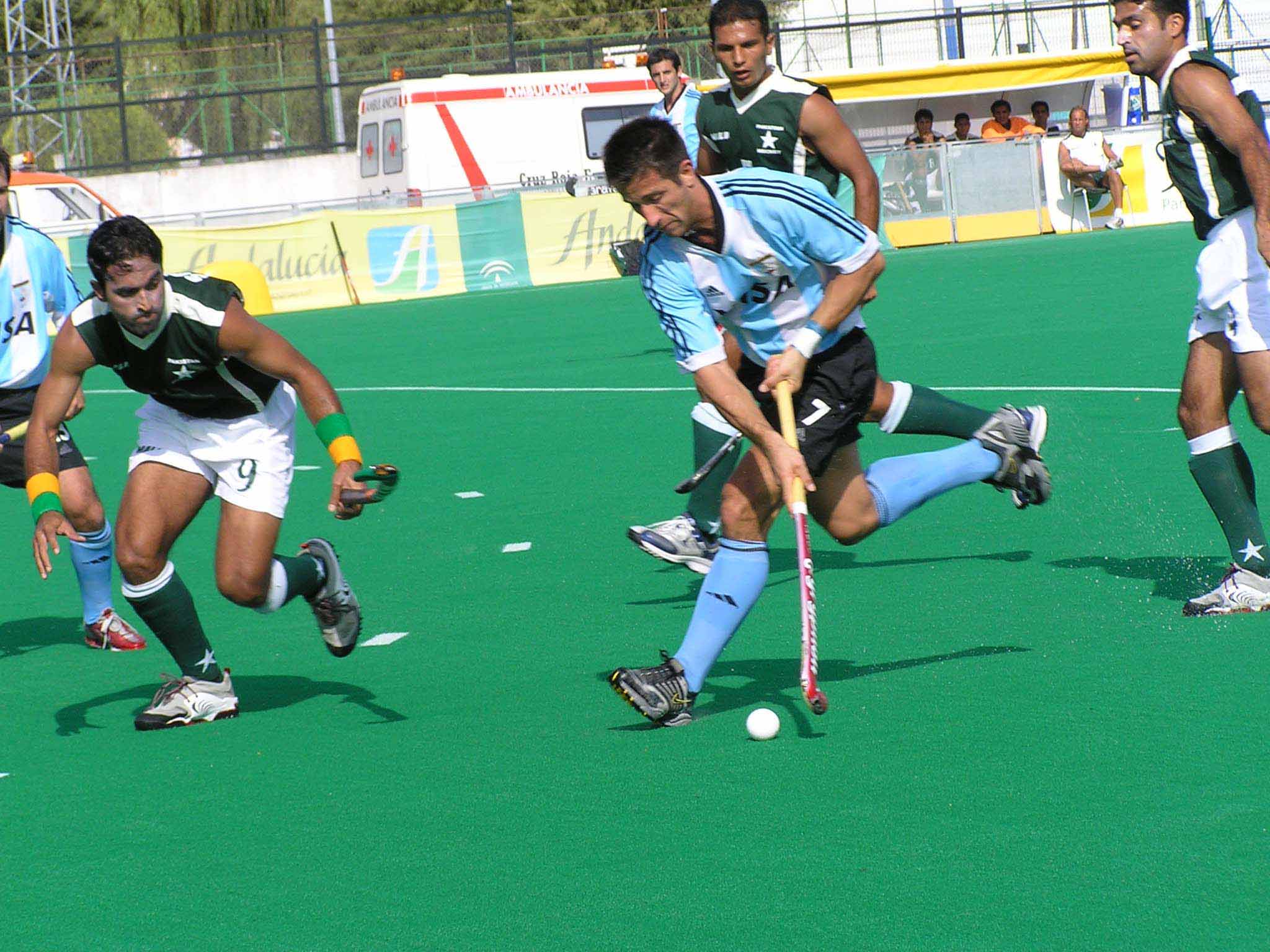 Pakistan field hockey players in green shirts contesting the ball with an Argentina player on a blue pitch