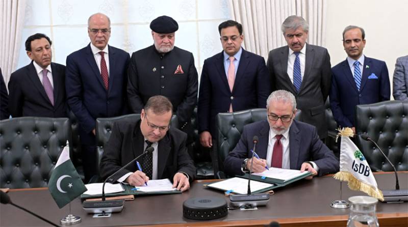 Two officials signing documents at a ceremony, with the Pakistan and Islamic Development Bank flags on the table in front of them.