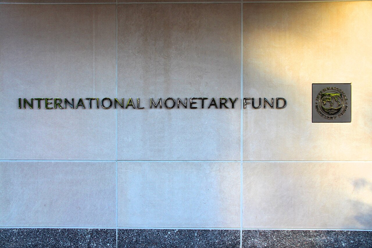 Street-level view of the International Monetary Fund headquarters in Washington, D.C., a large stone office building with a colonnade of tall pillars and rows of windows under a clear sky.