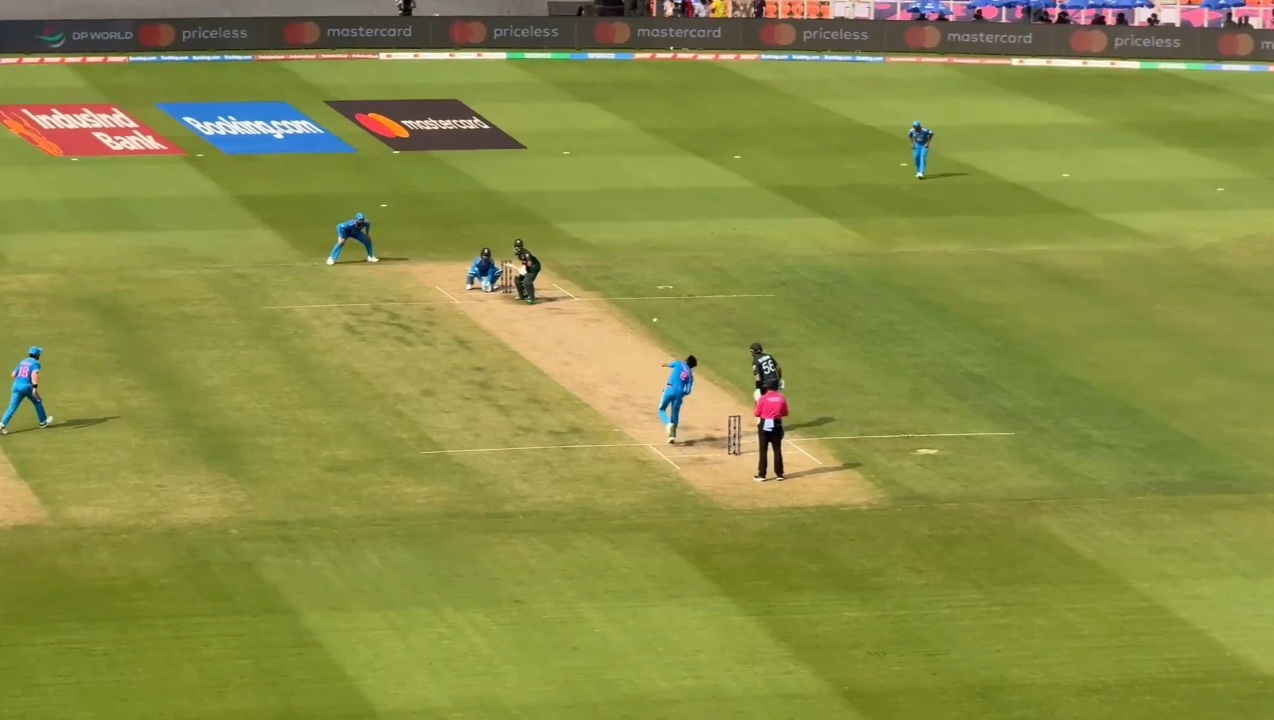 Cricket match in progress on a green pitch, with fielders in blue spread across the ground and a batter and wicketkeeper at the crease