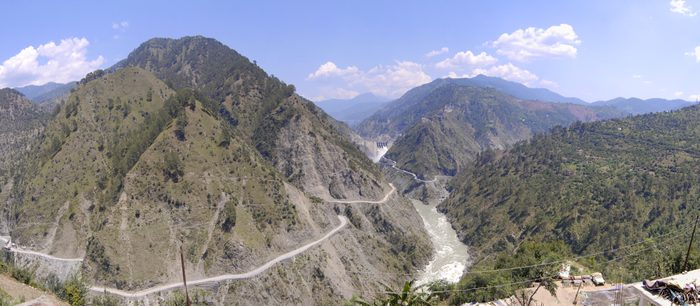 A river gorge seen from a hillside, with a winding road cut into steep, sparsely forested slopes above a pale green river, and mountains receding under a blue sky with scattered clouds.