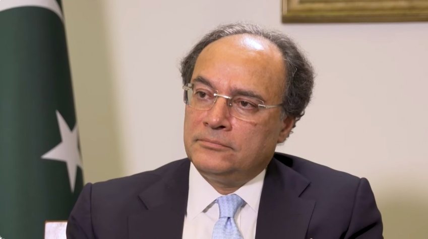 A man in a dark suit and pale blue tie seated in front of a green-and-white Pakistan flag