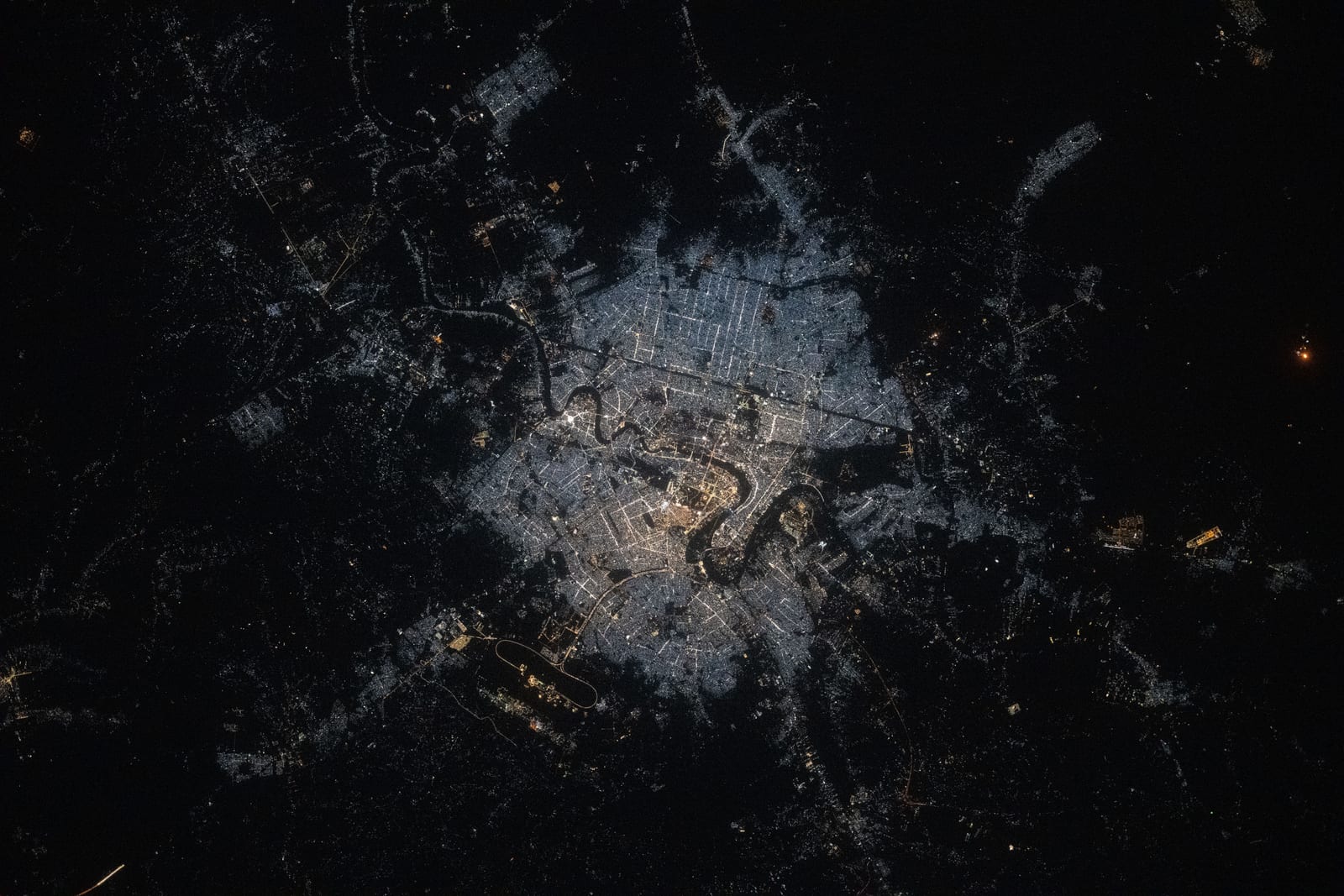 A night-time satellite view of Baghdad, with a bright grid of city lights divided by the dark ribbon of the Tigris River.