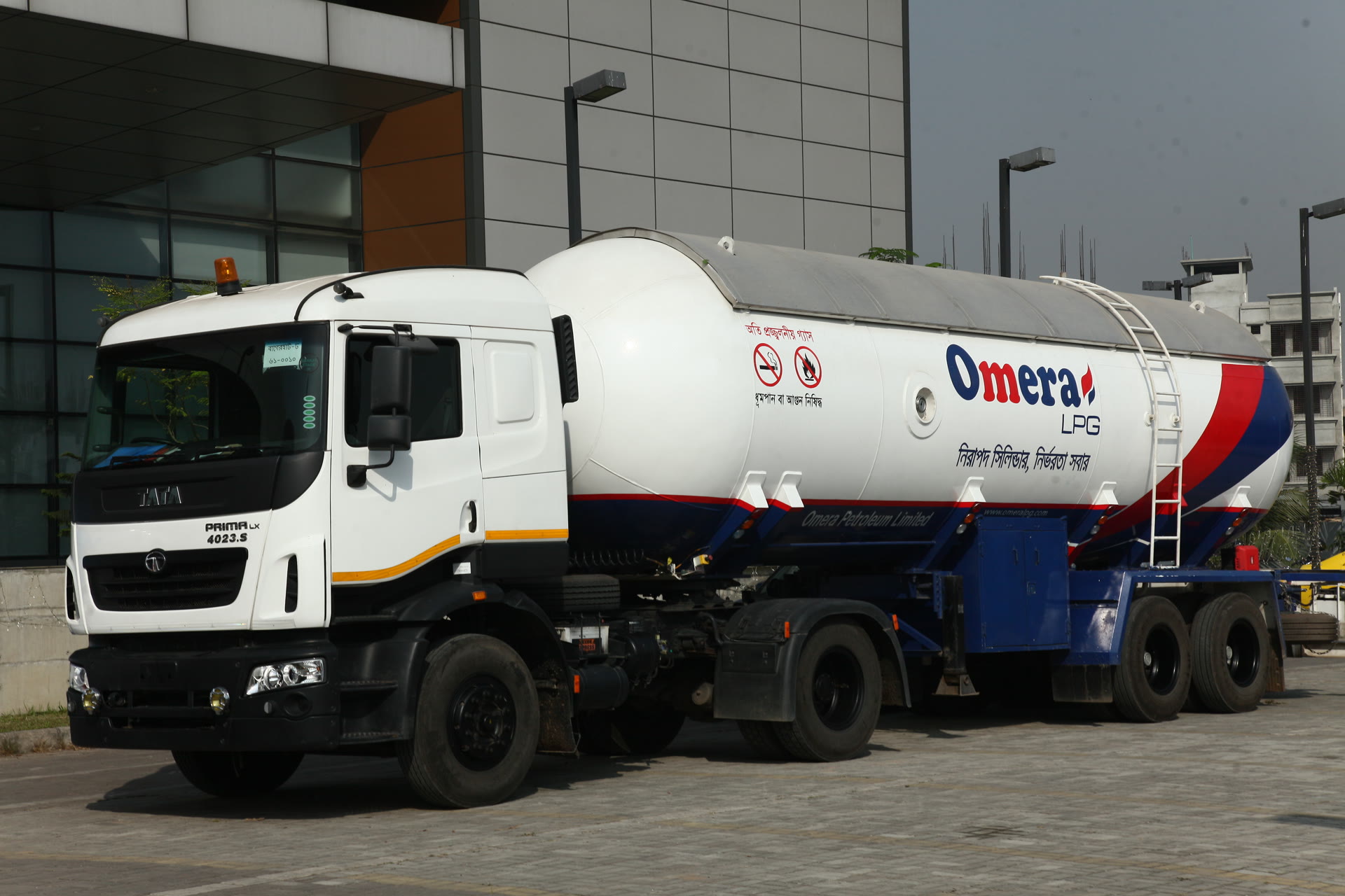 A white bulk liquefied petroleum gas road tanker with a long cylindrical tank marked with the Omera LPG name in English and Bengali lettering, parked on a paved forecourt beside a glass-fronted multi-storey building.