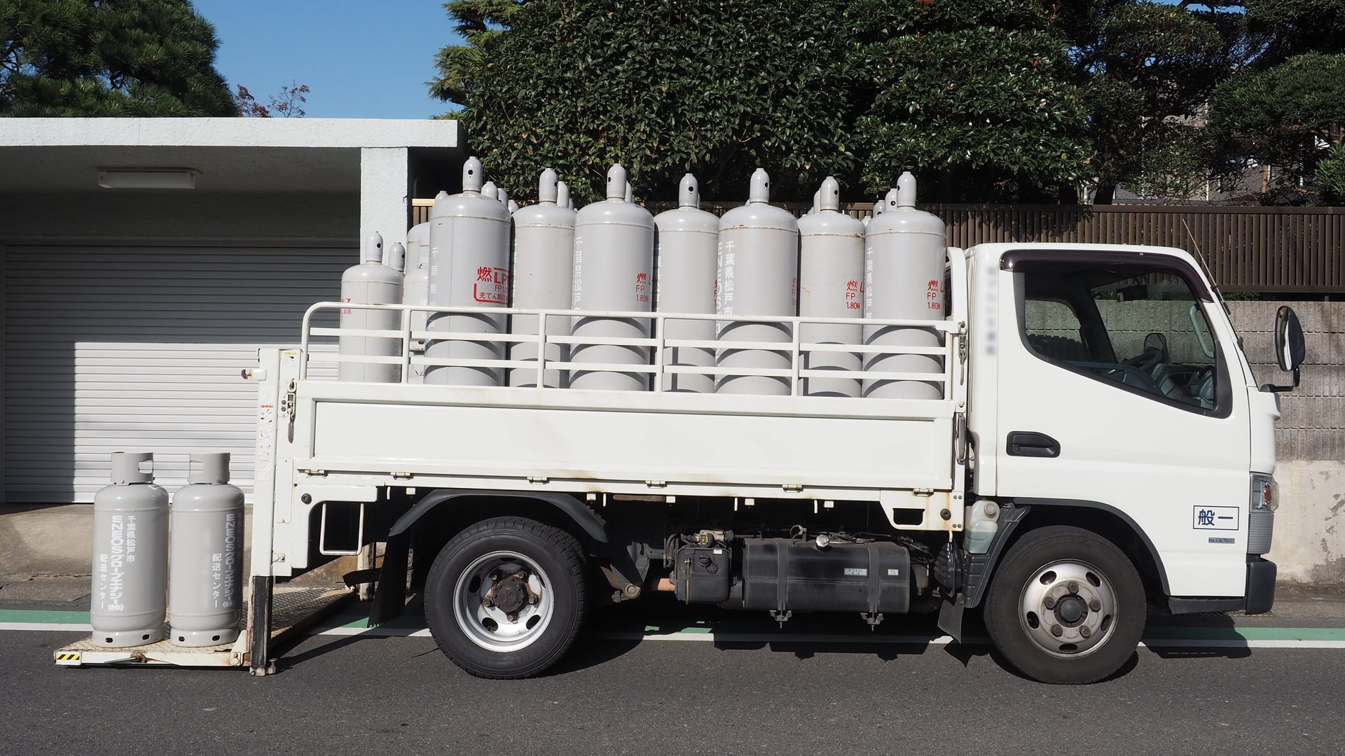 Household-size refillable LPG cylinders stacked in the bed of a white delivery truck, with two spare cylinders standing on the pavement beside it.