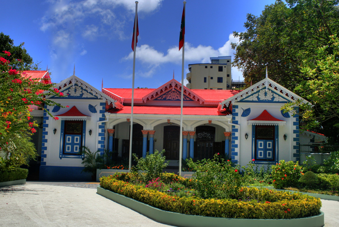 The Muliaage, the cream-white presidential palace on the Malé waterfront of the Maldives, photographed from the street with a black iron fence and tropical palms in front.