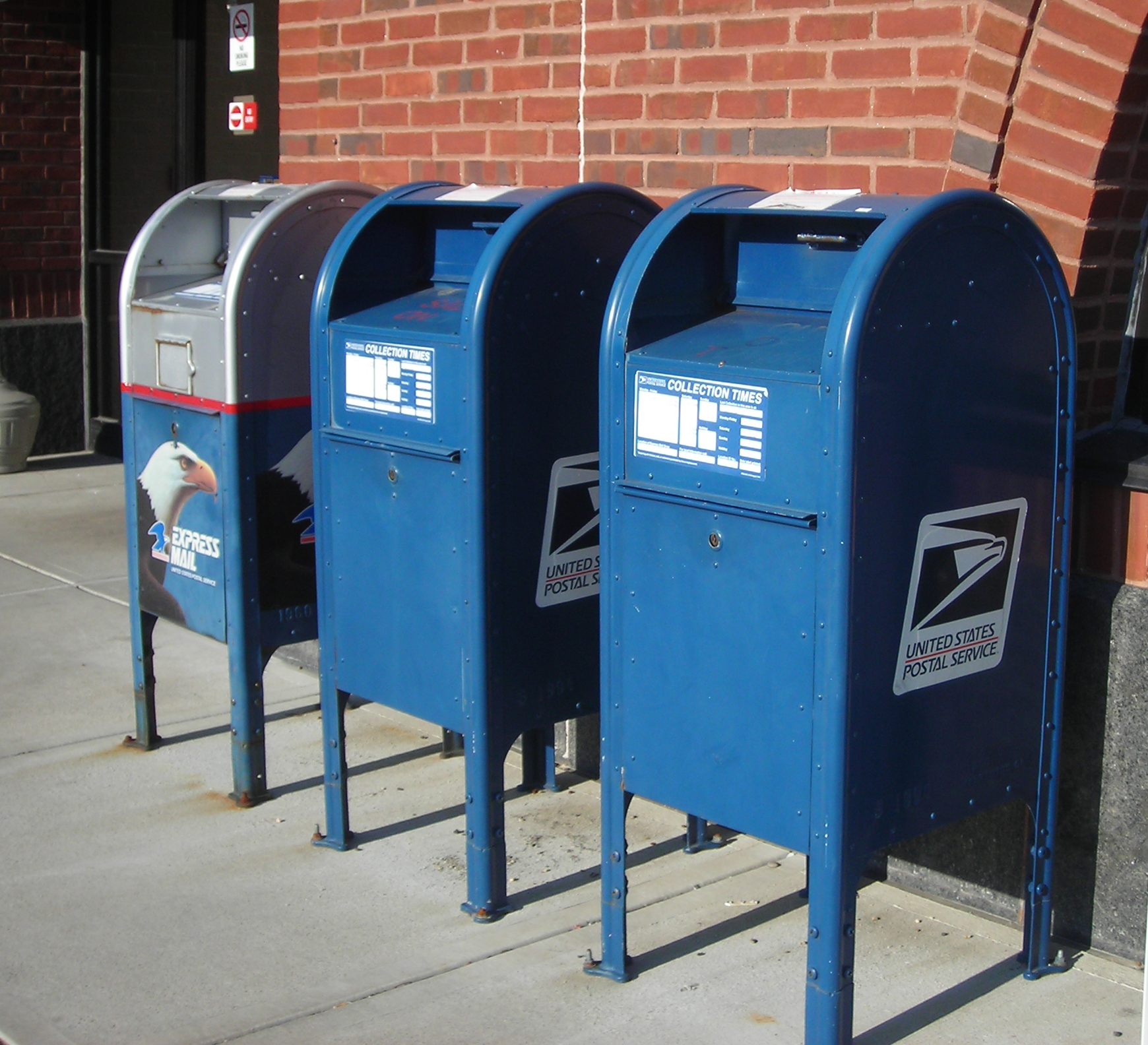 Three blue US Postal Service collection boxes lined up against a brick wall