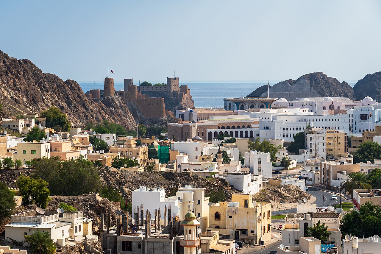 A daytime view of the Old Muscat waterfront in Oman, showing low-rise traditional buildings in the foreground, the Al Alam palace complex behind a row of stone fortifications, and turquoise water and low rocky hills in the background.