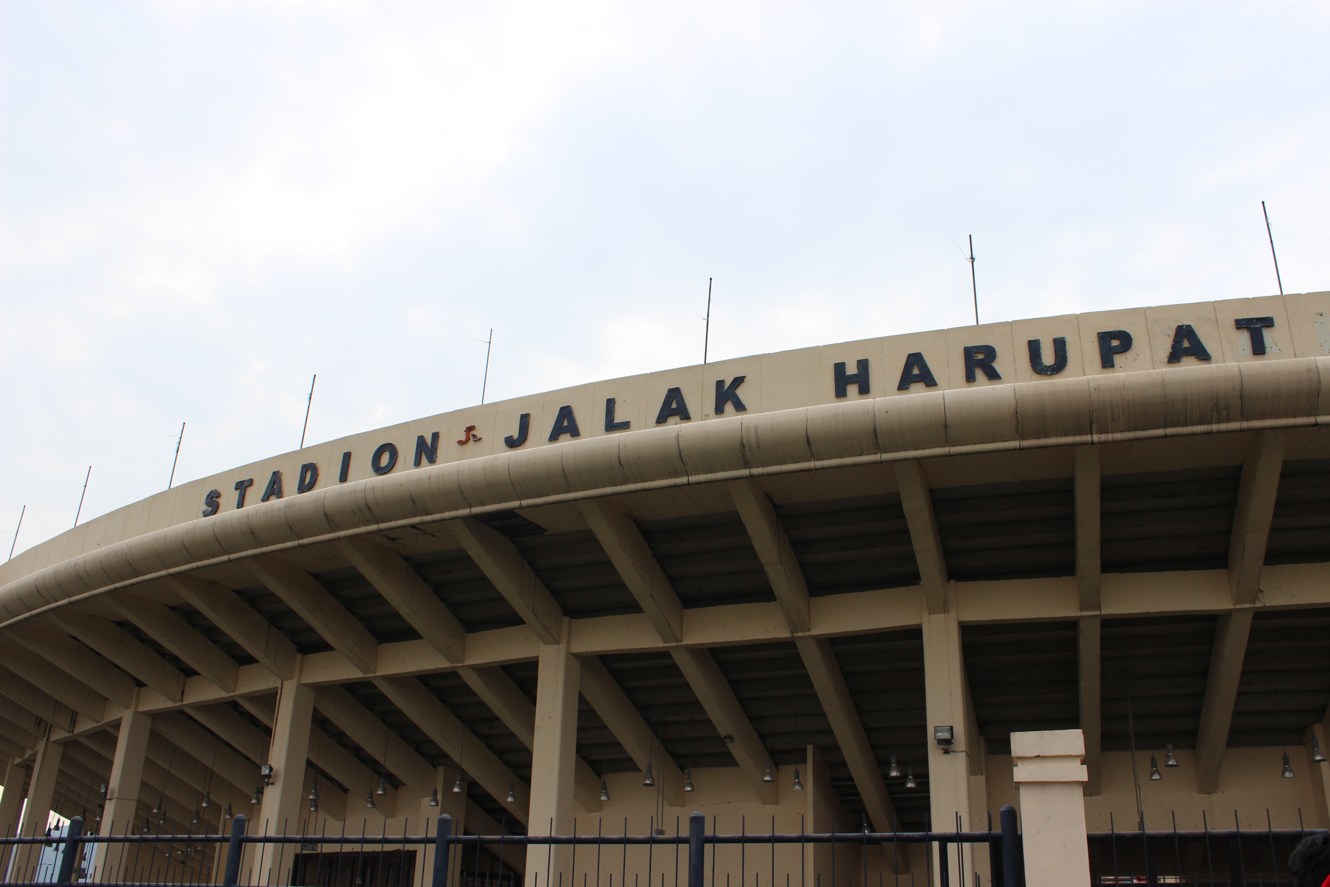 Exterior of Si Jalak Harupat Stadium in Bandung, Indonesia, with the stadium name mounted along its curved roof and concrete support pillars below