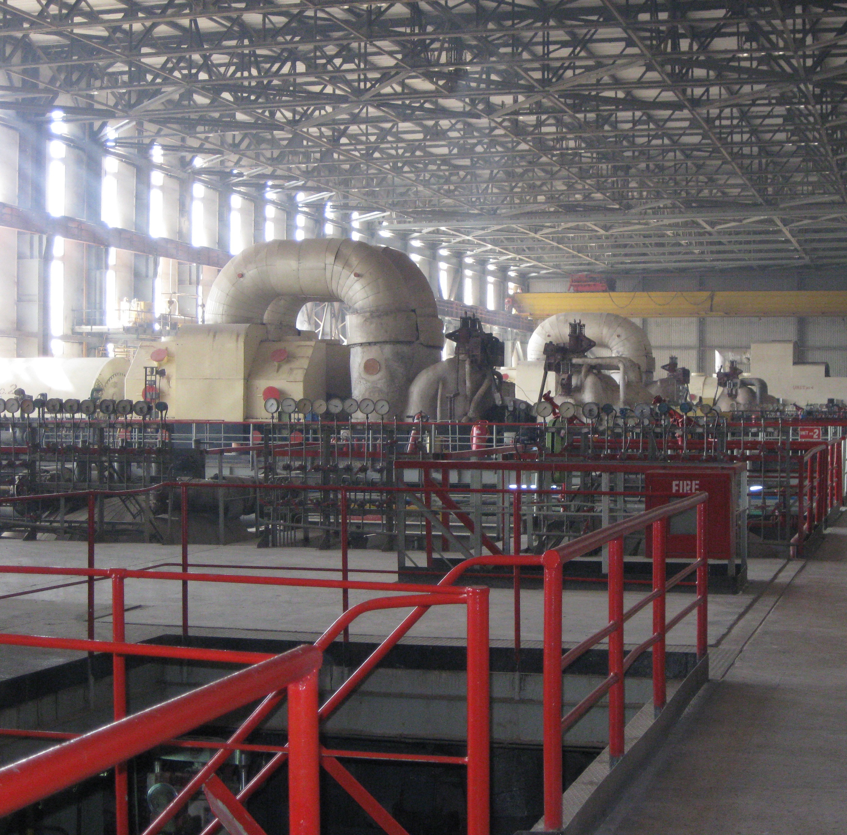 A large steam turbine and connected pipework inside the turbine hall of a thermal power station, with red safety railings in the foreground.