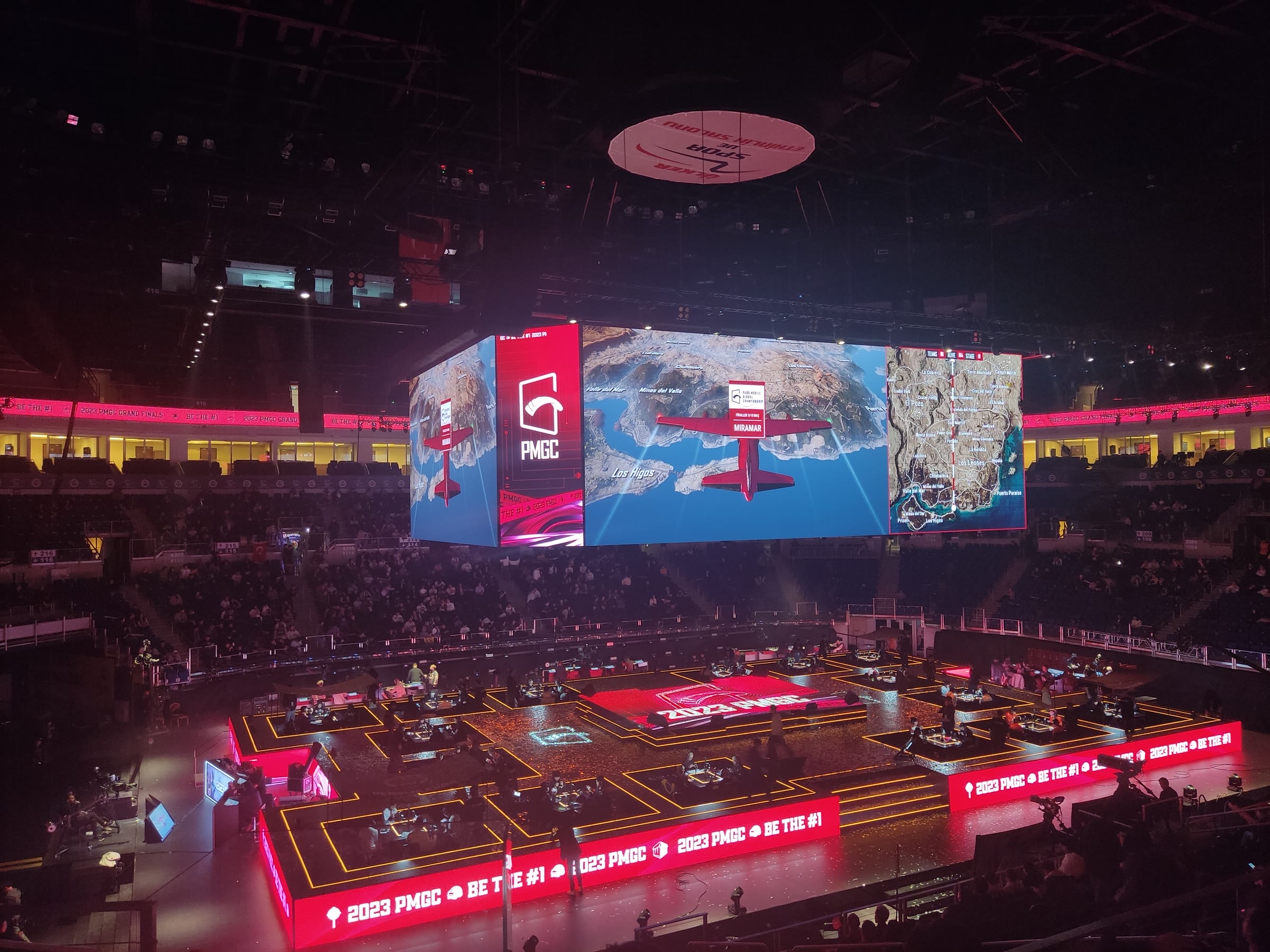 A darkened esports arena seen from the stands, with a large central screen showing a PUBG Mobile map above a red-lit stage ringed by player stations