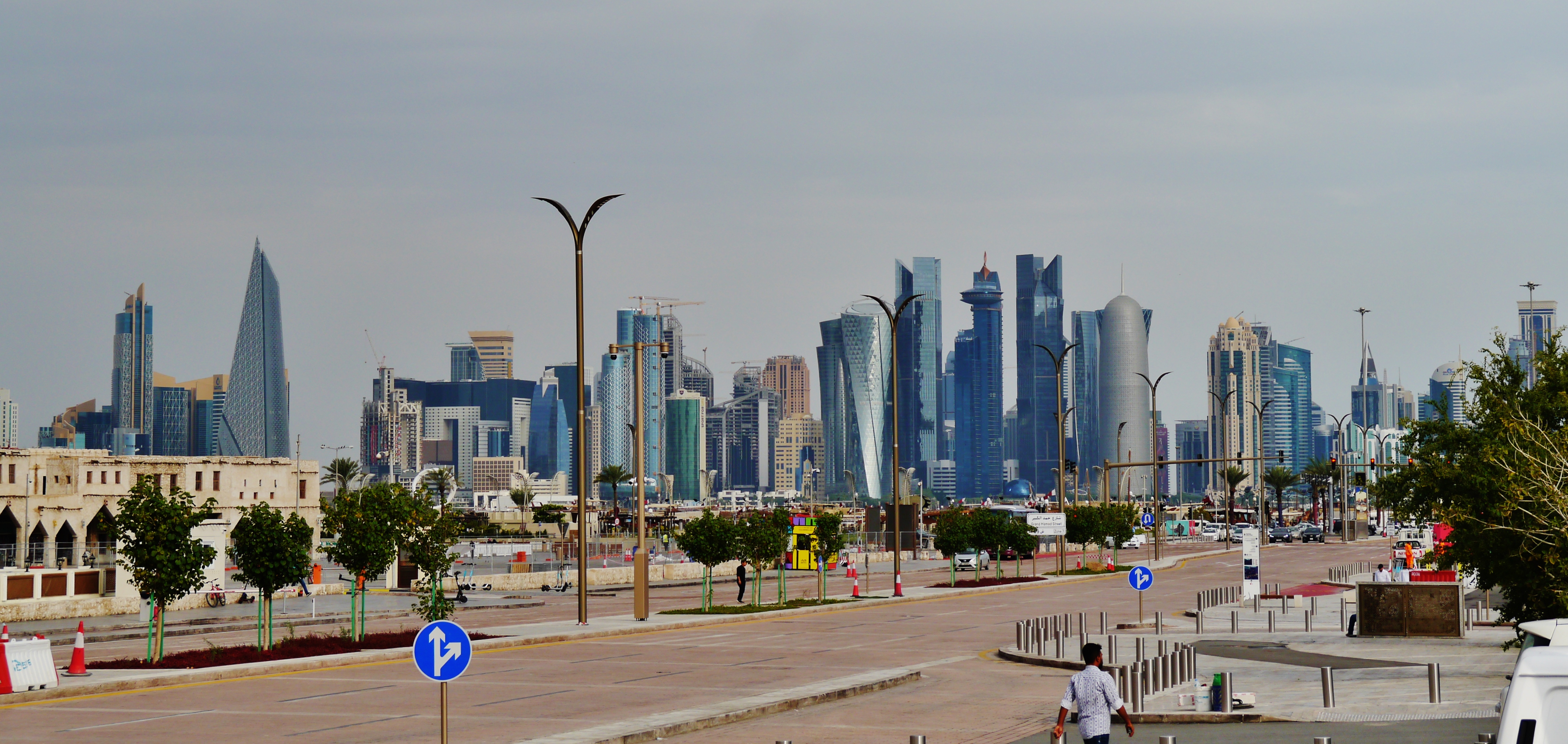 Doha's skyline with high-rise towers behind a wide waterfront road, street lamps, trees and traffic signs in the foreground.