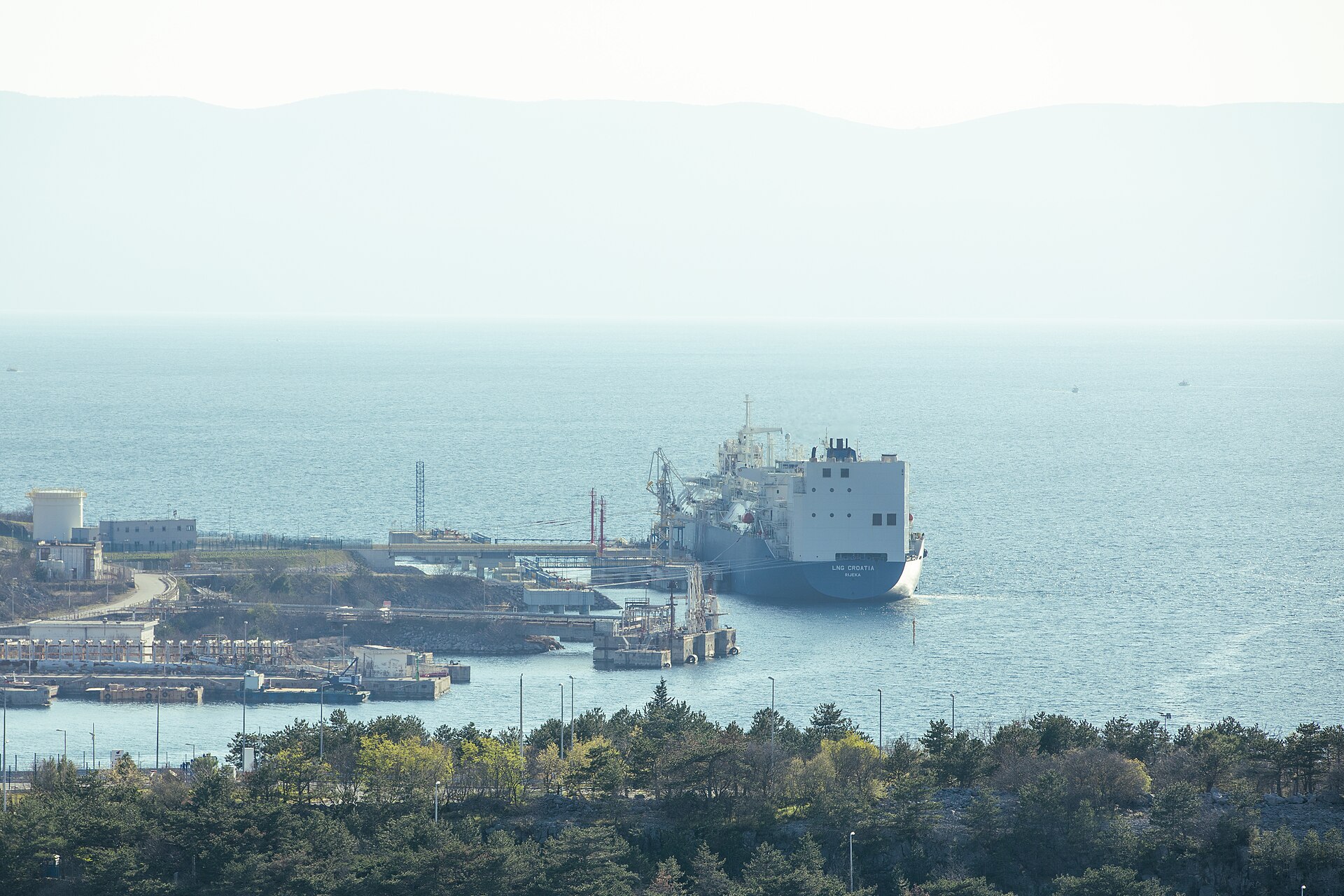 A large red-and-black tanker seen from above on open blue sea, with a distant coastline and terminal structures behind it.