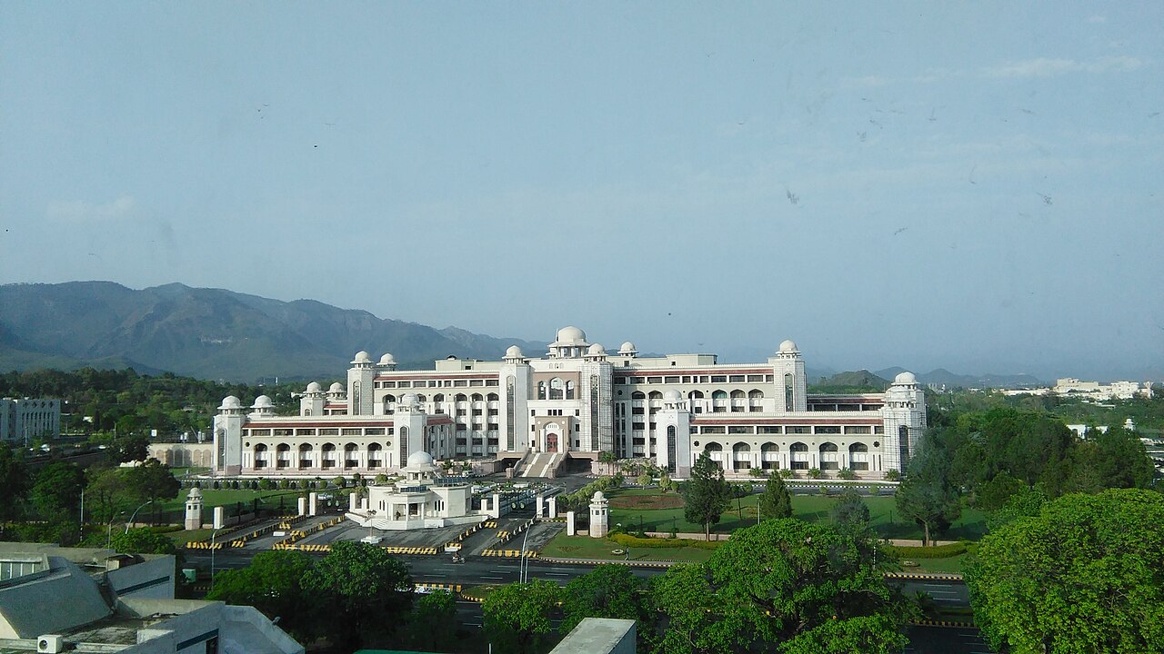 The white Prime Minister House complex in Islamabad, with domed roofs and arched facades, seen from a distance against the Margalla hills.