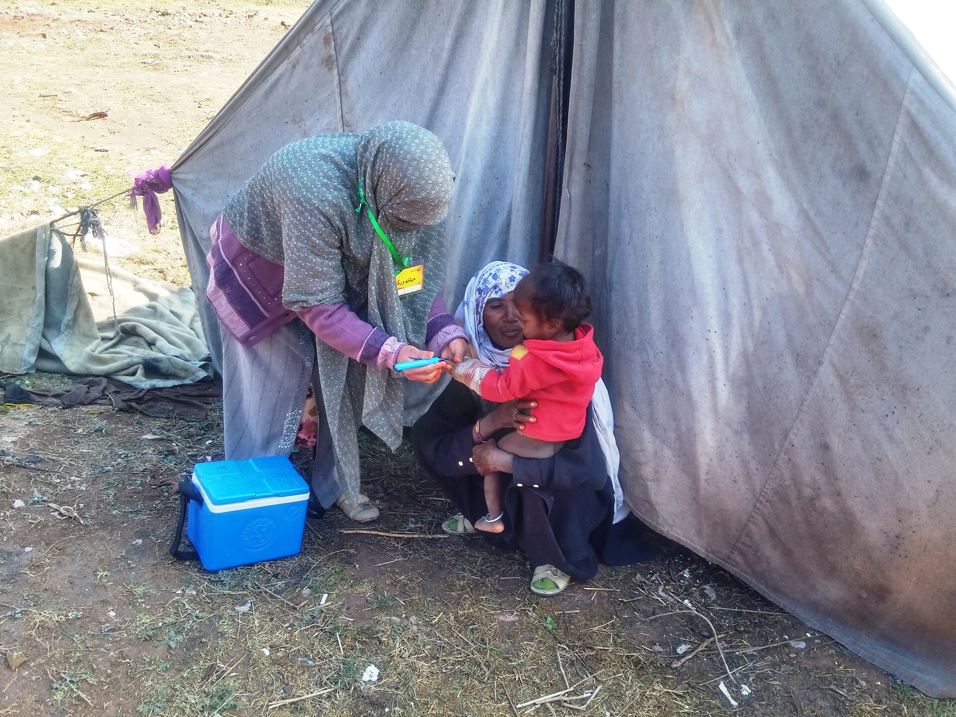 Health workers in a rural Pakistani setting administer oral polio vaccine drops to a young child held by an adult.
