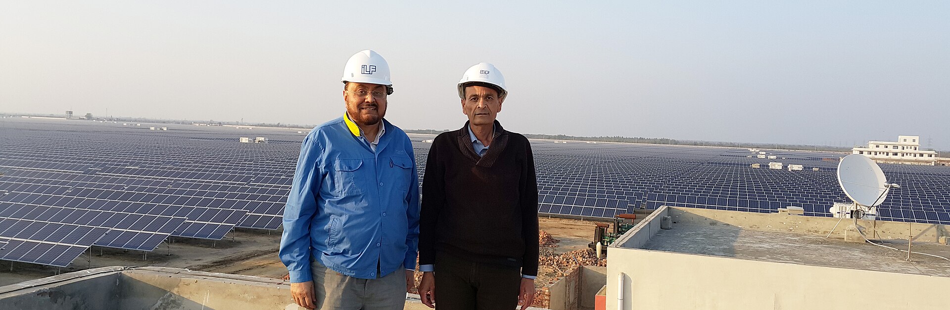 Two men in hard hats stand on a rooftop in the foreground with the rows of solar panels of the Quaid-e-Azam Solar Park stretching to the horizon behind them.